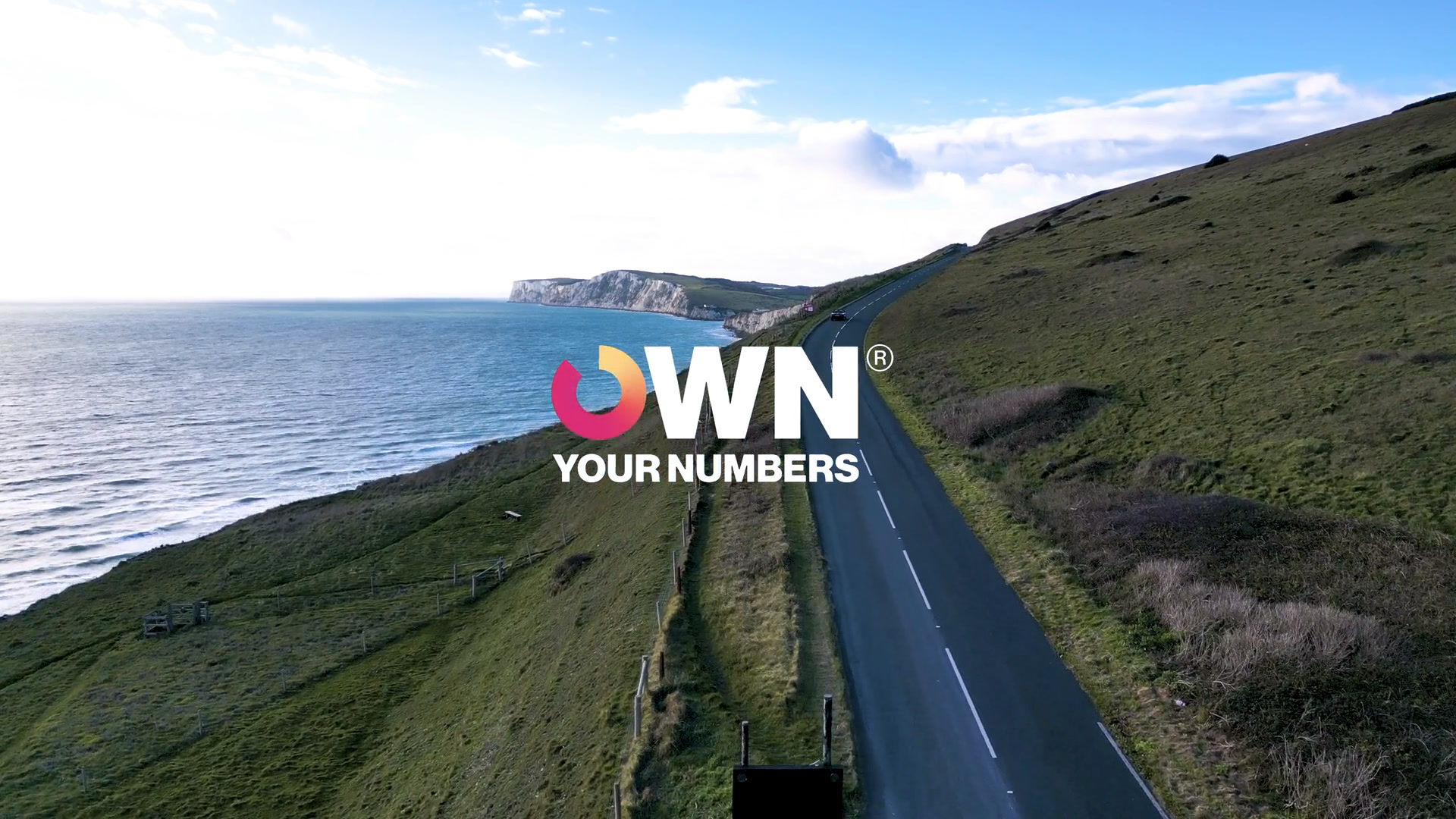 Drone still: the Military Road along the Isle of Wight coast, with the Own Your Numbers logo.